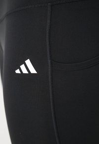 Close-up of black athletic leggings showing side pocket and white Adidas logo on thigh with visible seam stitching.