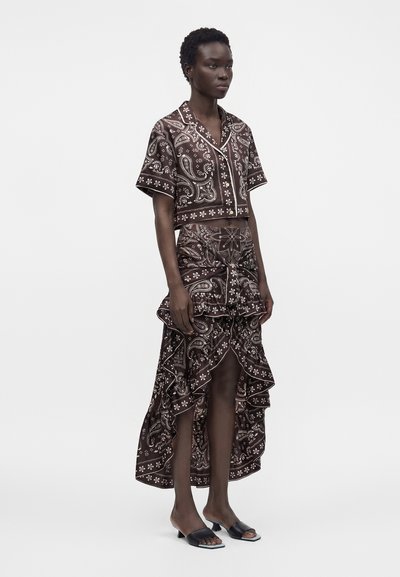 Femme portant une chemise courte à manches courtes imprimée paisley foncée assortie et une jupe asymétrique à volants avec des talons ouverts noirs, debout de côté.