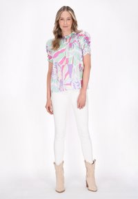 Farverig bluse med pastelfarvede mønstre i pink, grøn og lavendel, med puffede ærmer og en samlet halsudskæring, parret med hvide bukser.