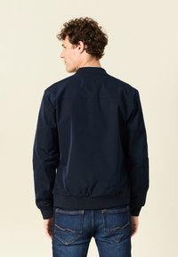 Chaqueta de bomber de color navy con cuello y puños acanalados, que presenta un tejido suave y ligero, y un cierre de cremallera. Llevada sobre unos jeans oscuros.