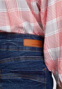 Punainen ja musta ruudullinen paita tummansinisillä farkuilla. Farkuissa on nahkalaatta, jossa lukee "TOM TAILOR Denim" ja eriväriset ompeleet.