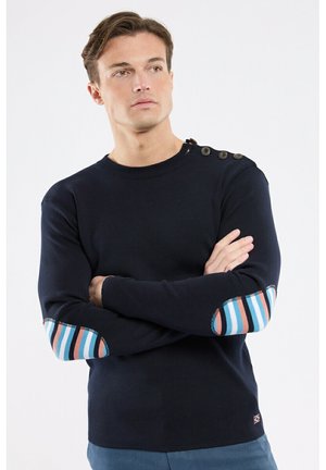Homme portant un pull noir avec des patchs de coude rayés colorés et des détails à boutons sur l'épaule, les bras croisés, regardant sur le côté.