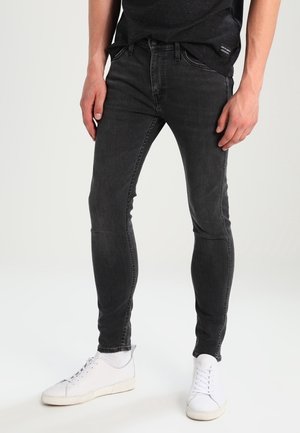 Mann trägt schwarze Slim-Fit-Jeans, schwarzes T-Shirt, weiße Socken und weiße Sneaker, steht vor einem einfarbigen hellen Hintergrund.