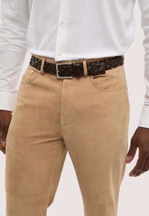 Uomo che indossa pantaloni in camoscio beige con tasche frontali, una camicia bianca con bottoni e una cintura intrecciata in pelle marrone scuro con fibbia d'argento.