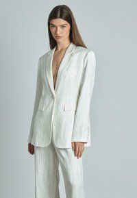 Witte blazer en bijpassende broek met een subtiel textuurpatroon. Beschikt over een diepe V-hals, twee zijzakken en knoopaccenten op de mouwen.