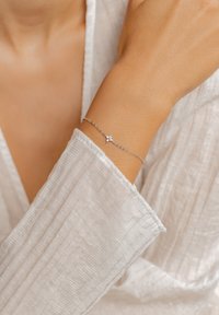 Bracelet en chaîne argentée avec un petit design de croix comportant trois pierres claires. Porté au poignet, mettant en avant une manche en tissu blanc texturé.