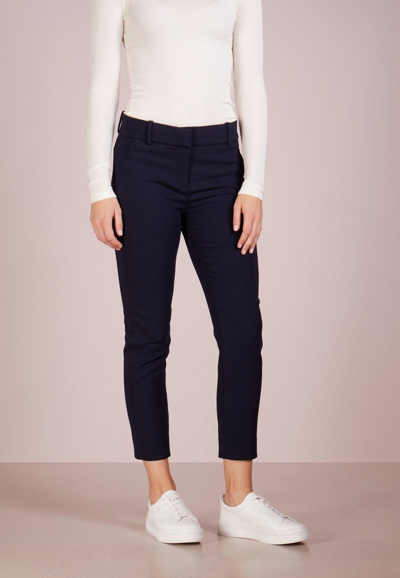 Femme portant un pantalon noir ajusté longueur cheville, un haut blanc à manches longues et des baskets blanches, debout devant un fond uni.