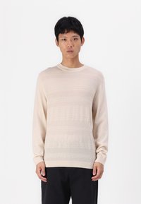 Bež pulover z dolgimi rokavi z teksturiranimi vodoravnimi črtami, okroglo ovratnik in rebrastimi manšetami. Tkanina se zdi mehka in lahka.