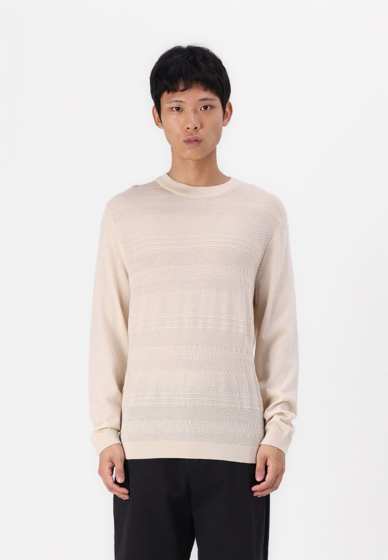 Bež pulover z dolgimi rokavi z teksturiranimi vodoravnimi črtami, okroglo ovratnik in rebrastimi manšetami. Tkanina se zdi mehka in lahka.