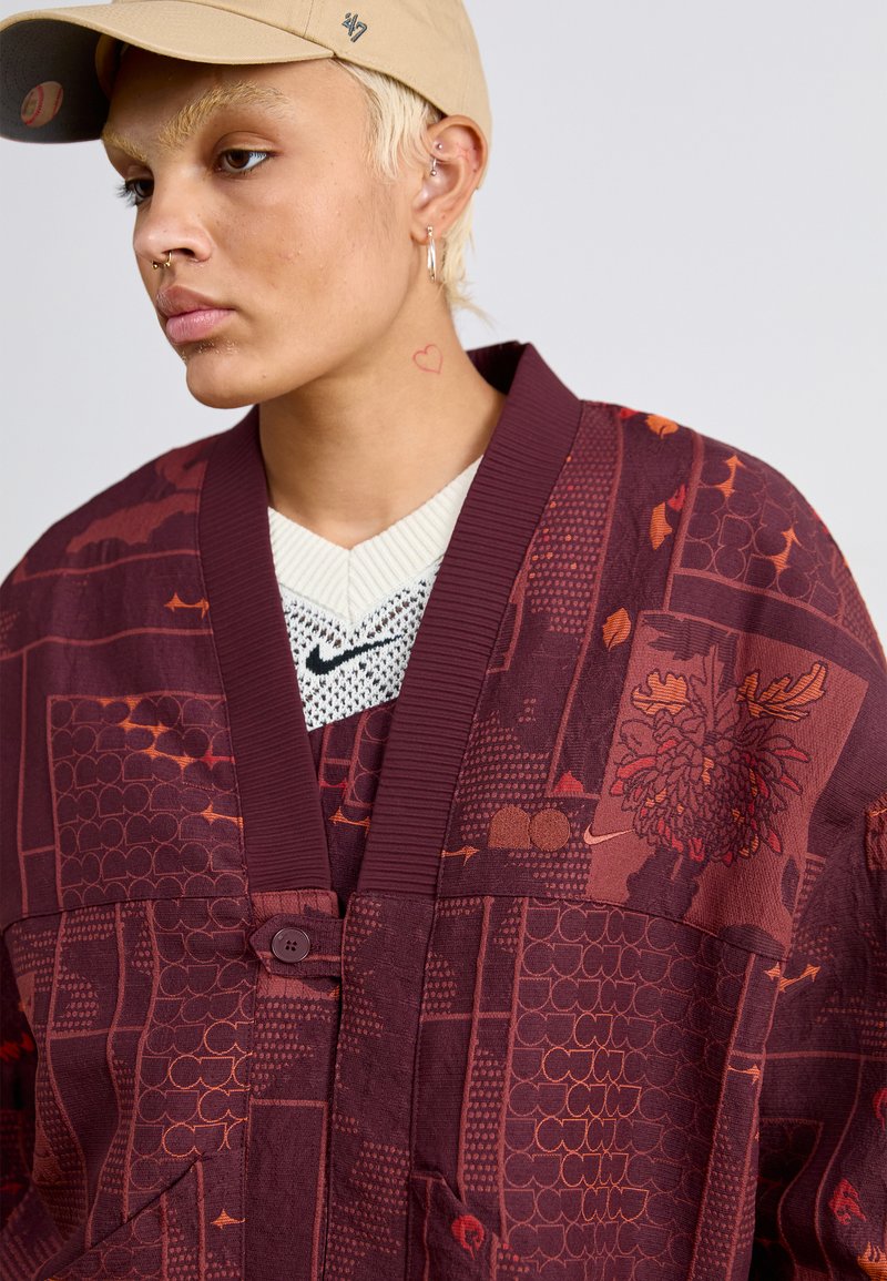 Weinrot gemusterte übergroße Jacke mit Knopfverschluss, die strukturierte Designs in Orange und komplizierte Grafiken aufweist. Darunter ein weißes Strickhemd.