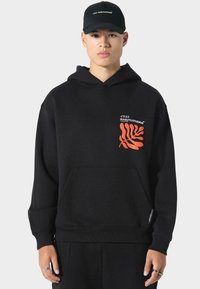 Černá mikina s kapucí a přední kapsou, zdobená červeným abstraktním vzorem a bílým logem na hrudi. Měkká tkanina s volným střihem.