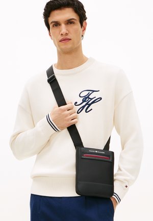 Μαύρη δερμάτινη τσάντα χιαστί με επίπεδο σχεδιασμό, με κόκκινη λωρίδα ως διακοσμητικό στοιχείο και λογότυπο στην κορυφή. Φορεμένη πάνω σε φόντο με ανοιχτόχρωμο πουλόβερ.