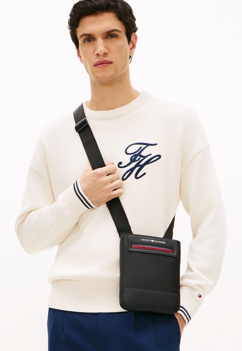 Μαύρη δερμάτινη τσάντα χιαστί με επίπεδο σχεδιασμό, με κόκκινη λωρίδα ως διακοσμητικό στοιχείο και λογότυπο στην κορυφή. Φορεμένη πάνω σε φόντο με ανοιχτόχρωμο πουλόβερ.