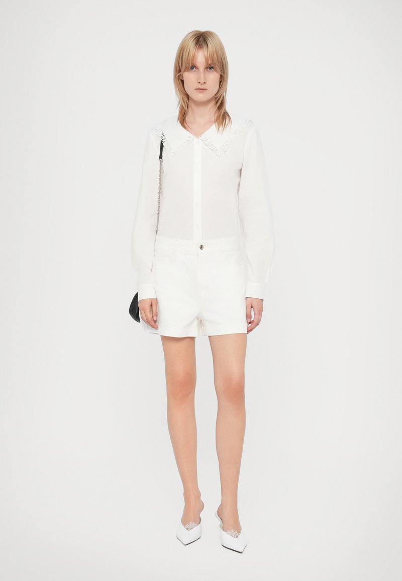 Chemise blanche à manches longues avec col en dentelle, associée à un short blanc. Le modèle tient un petit sac noir et porte des mocassins blancs avec une bordure en dentelle.