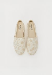 Des chaussures slip-on florales beiges avec un tissu texturé présentant des motifs en relief. Le logo de la marque est visible sur la semelle intérieure. Design à bout rond.