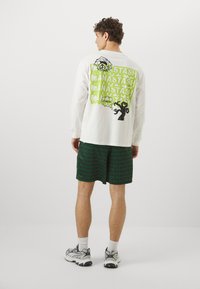 Langærmet hvid skjorte med grøn og sort grafik på ryggen, parret med grønne mønstrede shorts. Model iført sneakers og ankelstrømper.