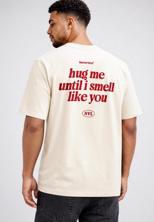 Mann mit kurzen lockigen Haaren, der ein hellbeiges T-Shirt mit rotem Text "hug me until i smell like you" auf dem Rücken trägt.