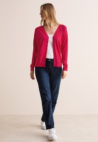 Cardigan rose vif en maille douce, doté d'un col V et d'une fermeture à boutons, associé à un jean bleu marine et des baskets blanches.