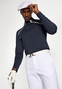 Camisa de golfe de manga longa azul-marinho com detalhes em branco, combinada com calças brancas e um boné branco. O modelo segura um taco de golfe enquanto usa uma luva branca.