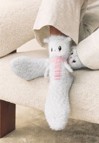 Calzini grigi pelosi con motivo di pupazzo di neve, con sciarpa rosa, occhi neri e finitura testurizzata, poggiati su un divano chiaro.