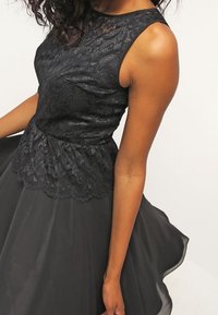 Svart klänning med en spetsöverdel och kjol i flätat satin. Toppen har ett texturerat mönster, medan kjolen har en slät finish och är lätt genomskinlig.