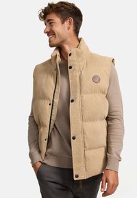 Beige Corduroy-Puffer-Weste mit hochgeschlossenem Kragen, schwarzen Druckknöpfen und einem Logo-Patch auf der linken Brust, getragen über einem hellbraunen Pullover.