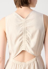 Beige sleeveless dress with a V-shaped back, featuring gathered detailing and a cut-out section near the waist, made from a smooth fabric.
