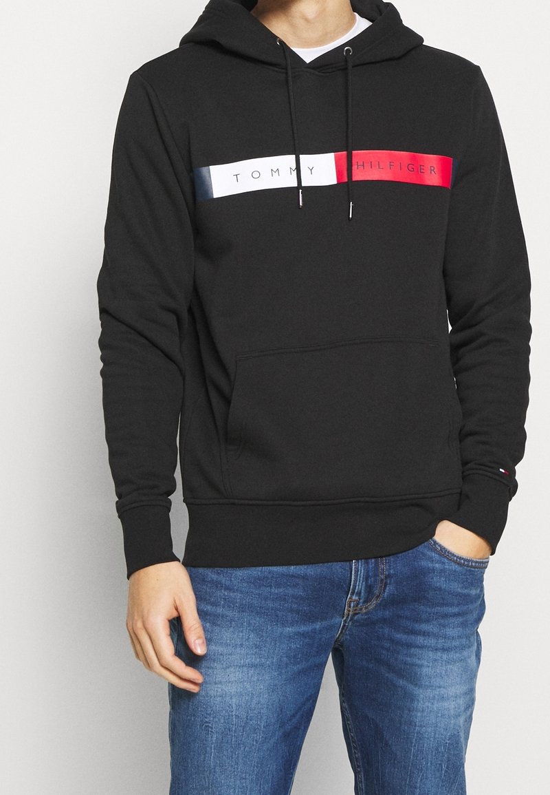 Felpa nera con tasca frontale, cordini sul cappuccio e una striscia orizzontale rossa, bianca e blu con il logo "TOMMY HILFIGER".
