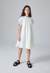 Hvid kjole med korte ærmer, sammenrynket overdel og broderet kant. Kombineret med sorte sandaler med en markant spænde.