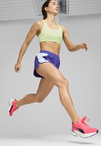 Tekstilni čevlji za tek v neonsko roza barvi z belimi podplati, v kombinaciji z svetlo zelenim športnim modrčkom in vijoličnimi kratkimi hlačami z svetlo modrimi detajli.