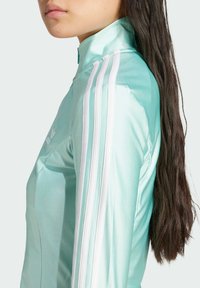 Chaqueta deportiva de color azul claro con cuello alto, que presenta detalles en franjas blancas a lo largo de las mangas y un logo sutil en el pecho. Textura suave.