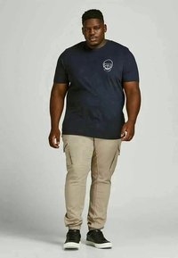 Homem de pé vestindo uma t-shirt preta com um logo de caveira branca, calças cargo bege e ténis pretos contra um fundo simples.