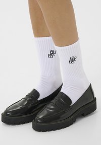 Črne usnjene loaferje z debelim podplatom, v kombinaciji z belimi rebrastimi nogavicami, ki imajo ob strani črn logotip.