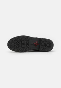 Suola di scarpa in gomma nera con un motivo di grip testurizzato, dotata di un logo centrale rosso. Il design enfatizza la durevolezza e la trazione.
