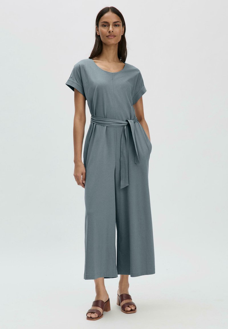 Hellgrauer Jumpsuit mit kurzen Ärmeln, tailliertem Gürtel, weitem Bein und seitlichen Taschen. Glattes Material mit lockerer Passform. Braune Absatzsandalen.