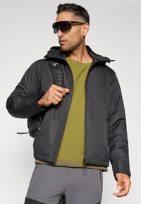 Veste noire avec capuche, dotée d'une fermeture éclair et de poches, portée sur une chemise verte texturée. Le mannequin porte des lunettes de soleil stylées de couleur sombre.