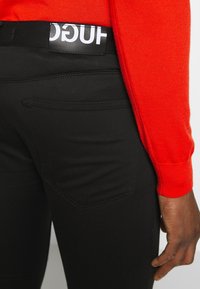 Persona con pantaloni neri e una cintura con il logo Hugo ben visibile, abbinata a una maglietta rossa a maniche lunghe, con la mano rilassata lungo il corpo.