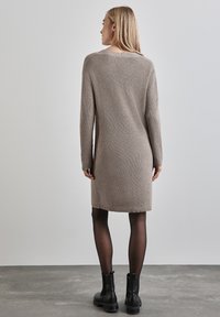 Beige geripptes Strickkleid mit langen Ärmeln, das über einen Rückenschlitz verfügt. Kombiniert mit schwarzen Ankle Boots und durchsichtigen schwarzen Strumpfhosen.