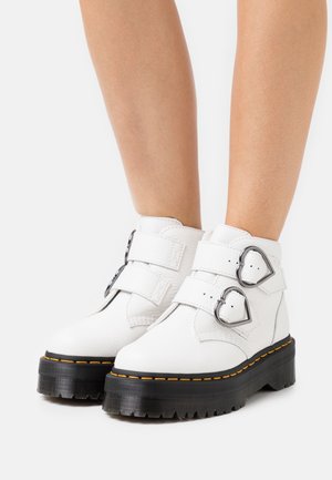 Botas de tobillo de cuero blanco con dos hebillas en forma de corazón, una suela negra gruesa y costuras amarillas contrastantes. Diseño plano con acabado texturizado.
