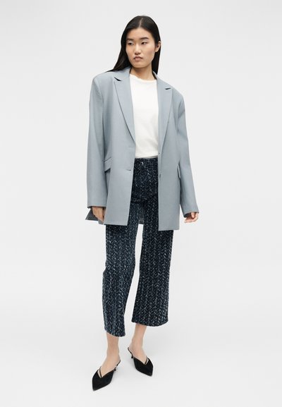 Femme aux longs cheveux noirs portant un blazer oversize gris clair, un haut blanc, un pantalon court à motifs et des chaussures slingback noires à bout pointu.