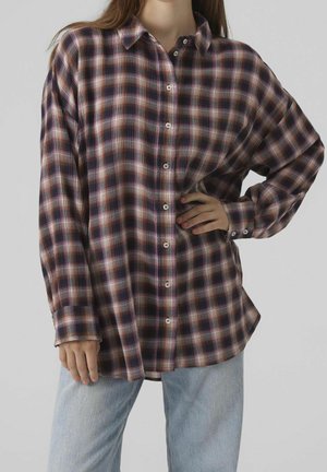 Kobieta ubrana w luźną, długą, zapinaną na guziki koszulę w kratę w odcieniach fioletu, czerni i brązu oraz jasnoniebieskie dżinsy.