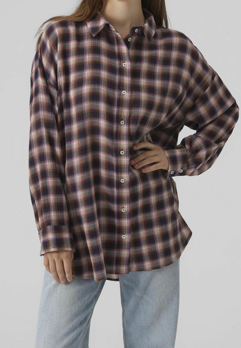 Femme portant une chemise à carreaux ample à manches longues et boutonnée, dans des tons violet, noir et marron, avec un jean bleu clair.
