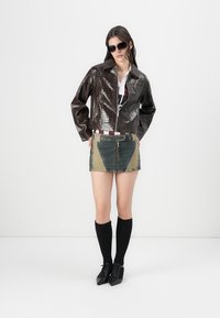Brun, glänsande konstläderjacka med en kortad design, tillsammans med en flerfärgad denimminikjol med lappar. Accentuerad med svarta knästrumpor.