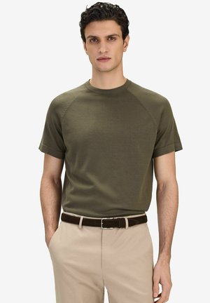 Mann steht mit den Händen in den Taschen, trägt ein olivgrünes Kurzarmhemd, beige Hose und einen dunkelbraunen Gürtel, vor einfachem Hintergrund.