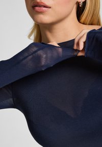 Vrouw in een marineblauwe sheer lange mouwtop die de stof bij de hals uitrekt, waardoor de semi-transparante textuur en de gouden oorringen zichtbaar zijn.