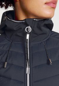 Giacca a vento nera con colletto alto, design a quadri, accessori in argento e coulisse. Texture liscia con dettagli lucidi lungo la cerniera.