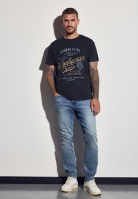 Černé bavlněné tričko s grafikou "Workwear Crew" ve zlaté a bílé barvě. Světlé modré džíny a béžové tenisky doplňují celkový vzhled. Upnutý střih, krátké rukávy.