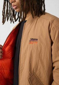 Bruine bomberjack met een oranje voering, ritssluiting en geborduurd "JOTT"-logo. De stof heeft een gladde, gestructureerde afwerking.