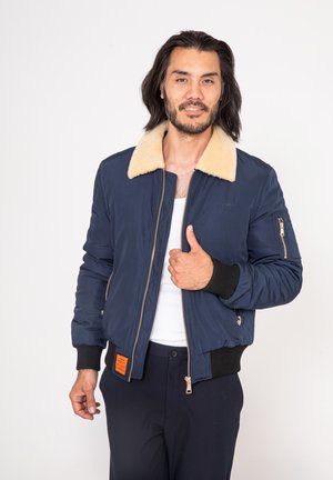Marine bommermantel met een beige faux fur kraag, ribbel manchetten en zoom, en een gouden rits; heeft een zijzak met oranje label detail.
