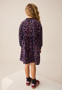 Robe violette à sequins avec manches longues, ornée de sequins multicolores. Portée avec des chaussettes foncées et des chaussures marron rougeâtre. Vue de dos affichée.